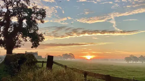 Serena Gough A sunrise image with a tree on the left, a fence running to the right hand bottom corner with sun run rising over a misty field with white clouds in a blue sky
