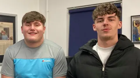 Two teenage boys standing next to each other and smiling. The boy on the left has light brown hair that is swept across his forehead. He is wearing a grey and blue t-shirt. On the right, the boy is wearing a white t-shirt and black puffer coat, he has short brown hair which is permed.