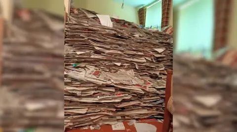 Making Space Piles of hundreds of newspapers stacked on top of each other to a height of around six foot. The top of a window with chequered curtains is barely visible on the right. 