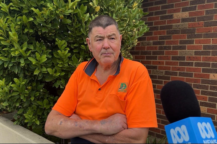 Man in fluro orange shirt with arms crossed and serious expression looks at reporter who is out of shot