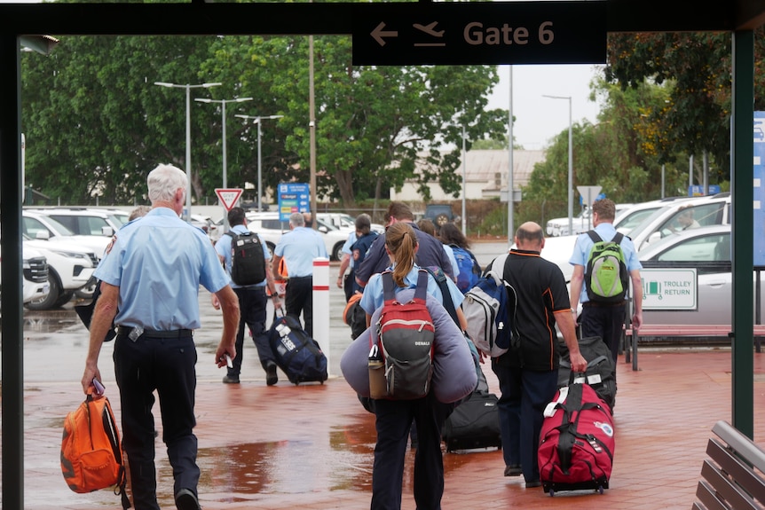 People carrying suitcases and bags walk away into a carpark at an airport on the wet ground.