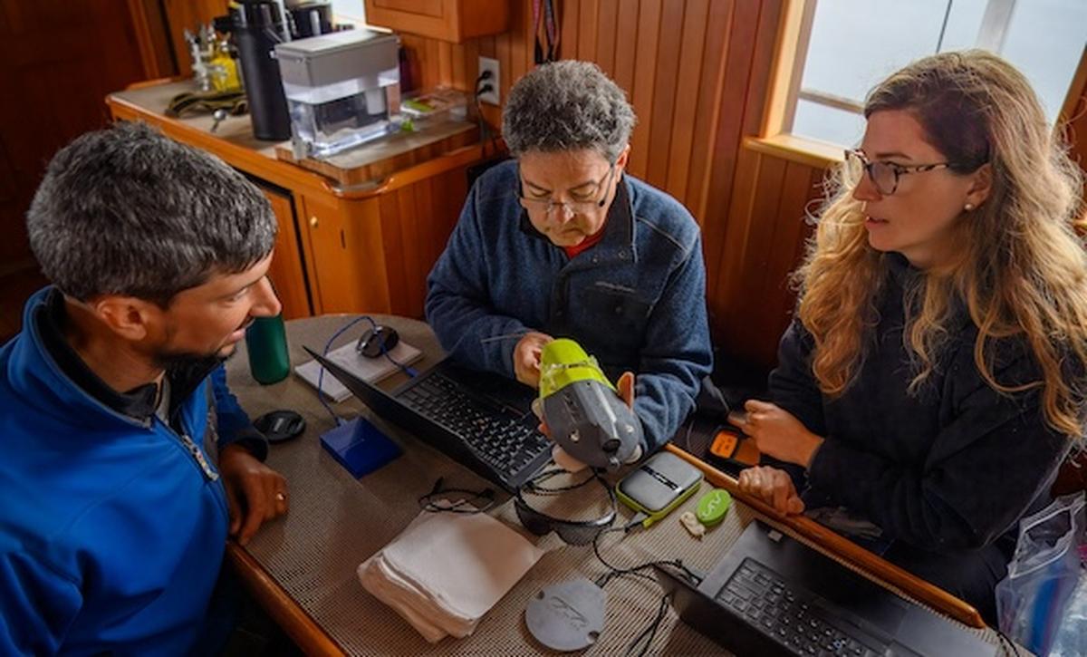 Researchers Mike deRoos (Cetacea Contracting, Ltd), Andrew Trites, and Sarah Fortune preparing gear for their research. The three are gathered around a small table on a boat. There are two laptops open on the table. The scientist in the center is working on a tracking device.