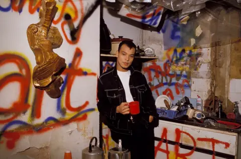 Redferns / Getty Images Roland Gift poses for a photograph while leaning against the wall of a dirty, delapidated kitchen with graffiti all over it and holds a mug while looking at the camera wearing a denim top, taken in 1980.