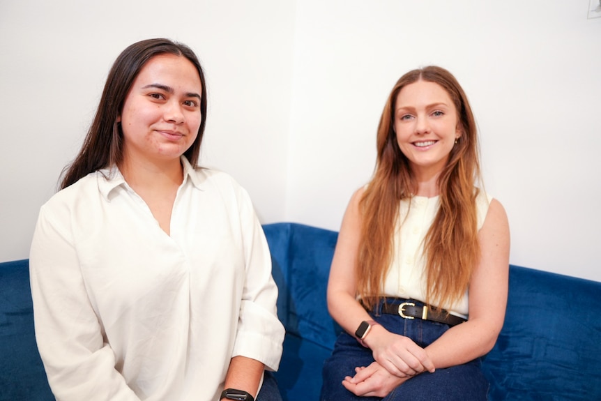Jemima left in a white top, Sonia right in a white top and jeans, smile while sitting on a blue couch.