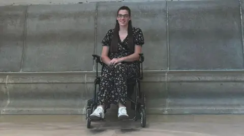 Supplied Hannah Dickerson wearing a black and floral patterned outfit while sitting in a wheelchair in front of a concrete wall. She is smiling and looking into the camera.