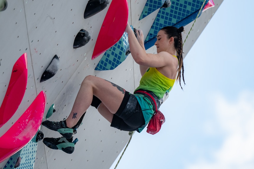 Climber Sarah Larcombe holds on to a steeply angled climbing wall. She wears a gold tank top and has her hair in plaits.