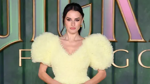 Getty Images Amber Davies wearing a v necked yellow dress with a style that highlights the use of more material on the shoulders. She is stood in front of Wicked signage suggesting she is at a movie premiere.