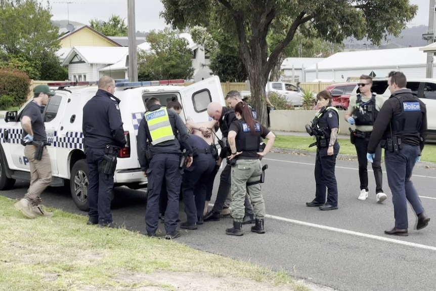 Nine police surround one man (face not shown) as two of them put him in the back of a divvy van in a residential street.