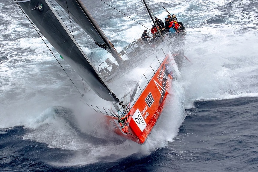 A large sail boat with crew in rough seas.