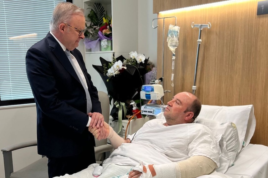 Prime Minister Anthony Albanese stands while holding Ahmed's hand while he lies in a hospital bed with his left arm in bandages.