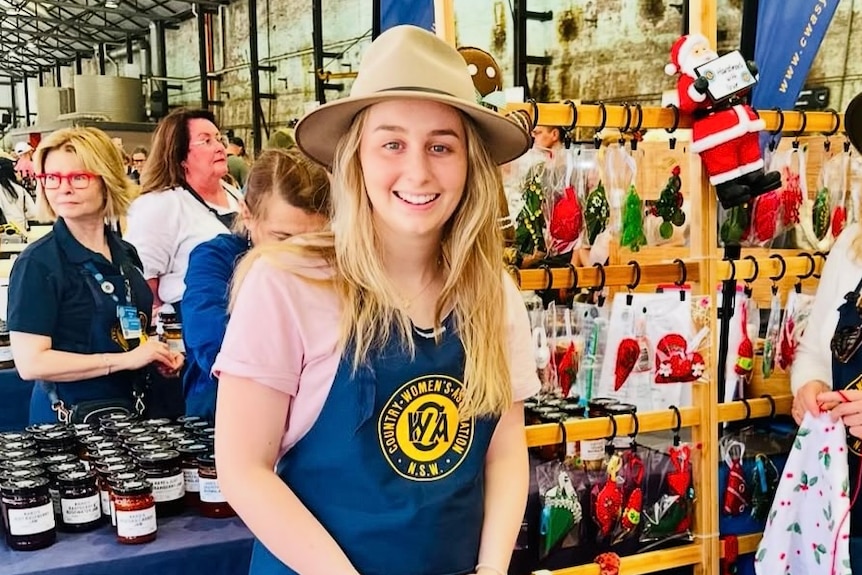 Maddy Langabeer left in a hat, smiling, wearing a CWA apron at a marketstall.