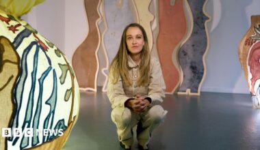 Bethany Williams wears a beige jacket and trousers, she has brown/ blonde hair and crouches in an exhibition which has colourful lights and arching coloured panels in the background.