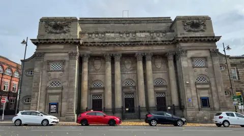 The vacant Queen's Theatre building in Burslem - a large, old theatre building. A number of cars are parked in front of it on the road.