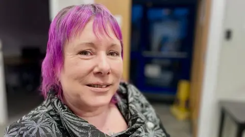 Mark Norman/BBC Leanne Nagel poses close to the camera. She has dyed purple hair and is wearing a black patterned blouse.