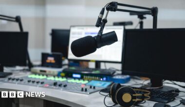 A radio studio with a mixing desk and a microphone and a set of headphones on the desk. There are three computer screens with the one in the middle switched on.