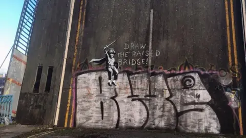 BBC A painted mural depicting a small boy is sitting on top of large letters wearing a cape and holding a wooden sword with a pencil stuck to the top of it. A caption next to the image reads "Draw the raised bridge!"
