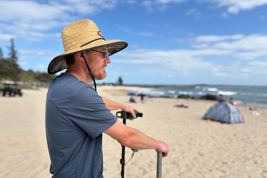 A man holding a metal detector looking at towards the ocean, at the beach.