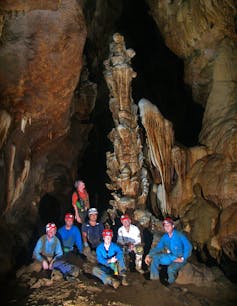 A group of 7 cavers in blue and white overalls and red helmets pose in front of an 8m high intricately decorated stalagmite in a dark cave.