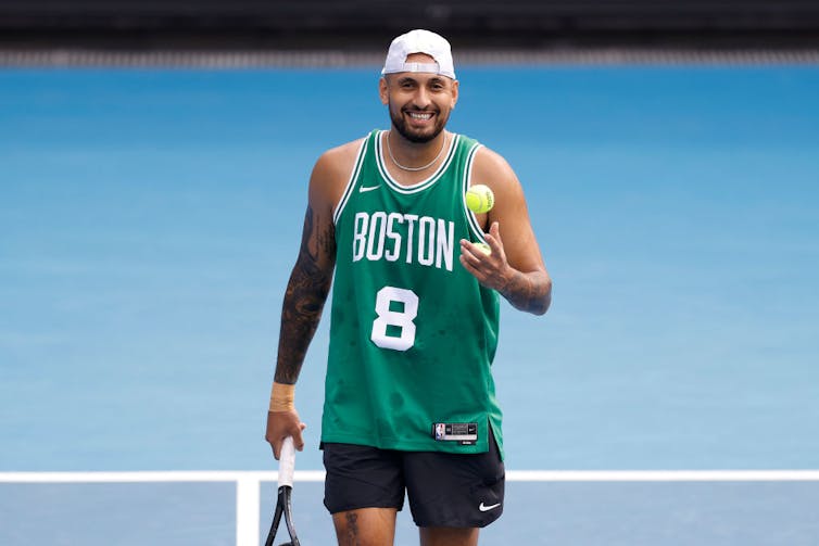 Young man smiling while holding a tennis racket and a tennis ball, wearing a green basketball jersey and a backward white cap.
