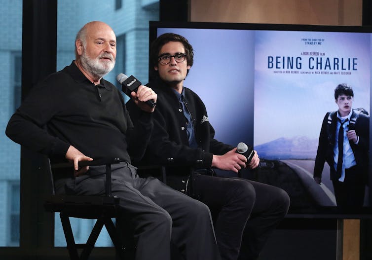 Rob Reiner holds a microphone next to a young man with a banner for the movie 'Being Charlie' visible in the background.