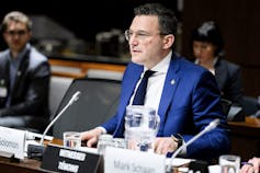 A man with dark hair in a blue suit sits behind a microphone.