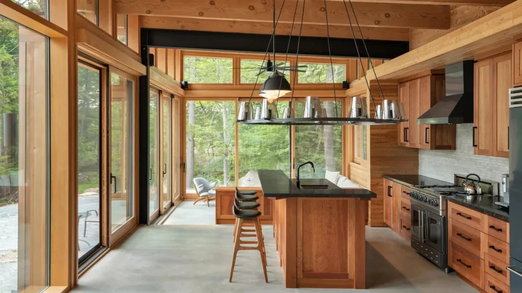 A light-filled kitchen surrounded by floor to ceiling windows that look out into the forest surrounding the lakehouse retreat, with concrete flooring and wooden beams. 