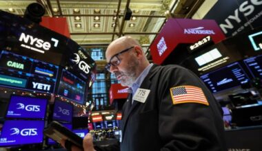 A trader in a jacket with a US flag patch works on the NYSE floor, surrounded by digital stock ticker screens.