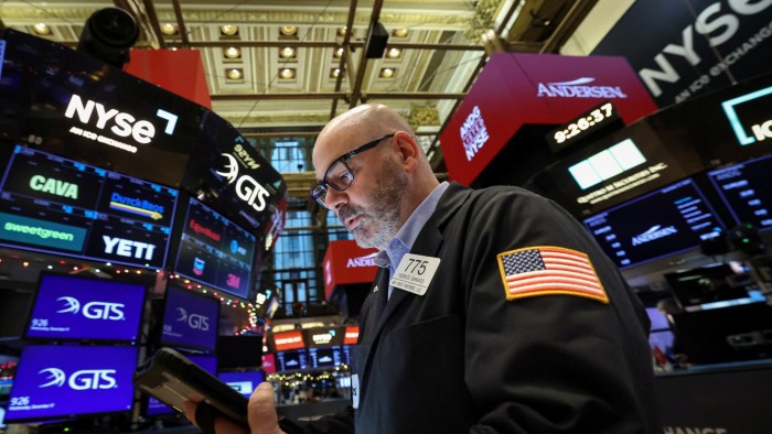 A trader in a jacket with a US flag patch works on the NYSE floor, surrounded by digital stock ticker screens.