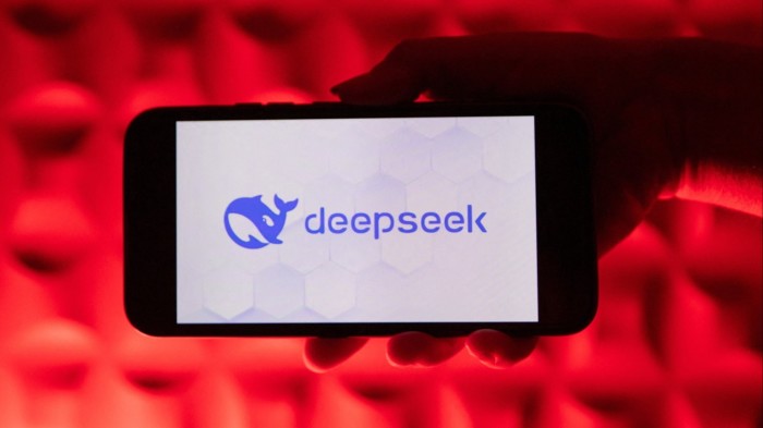 A hand holds a smartphone displaying the Deepseek AI logo against a vivid red illuminated background.