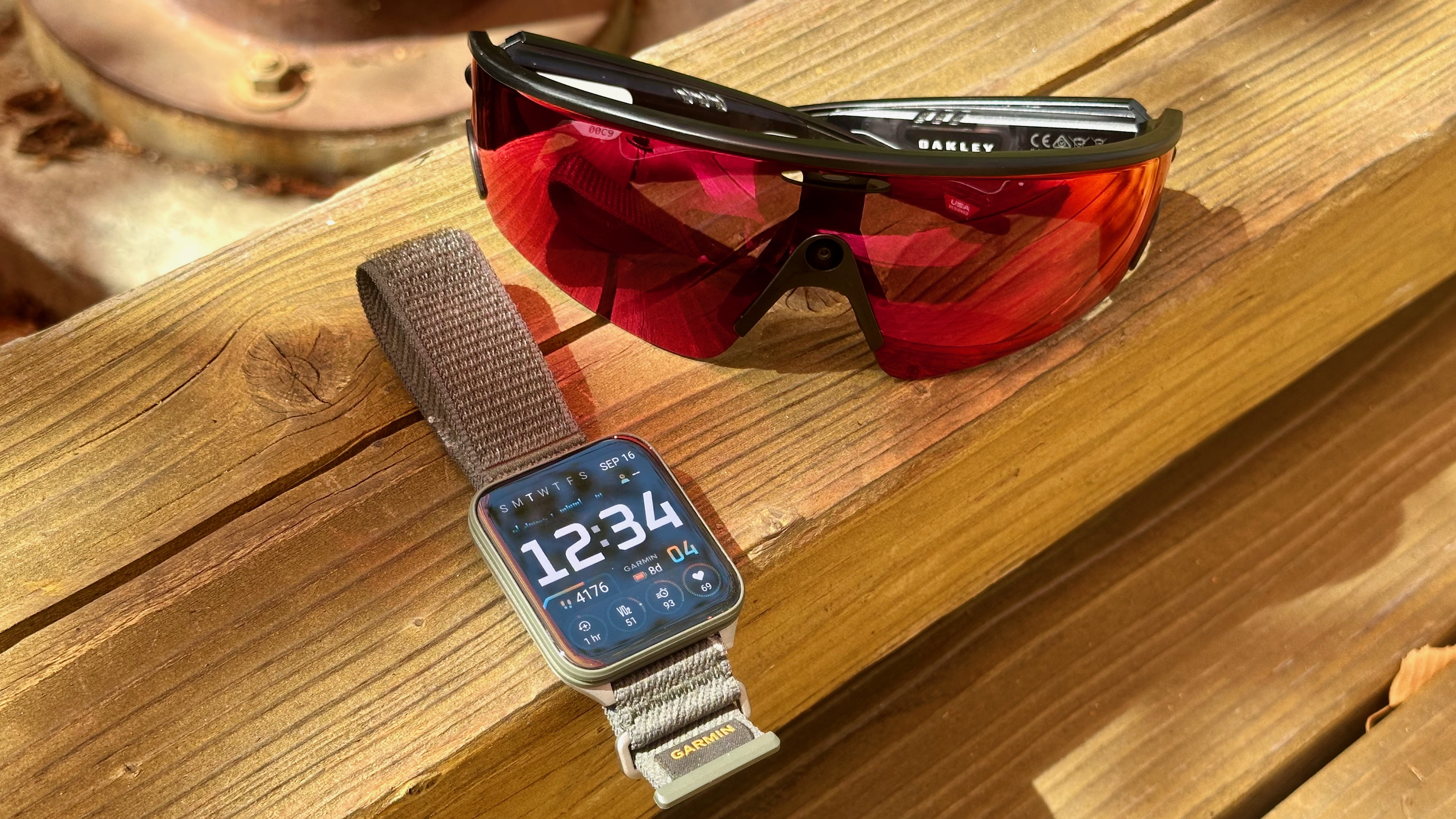 Photo of the Oakley Meta Vanguard smart glasses and Garmin Venu X1 smartwatch sitting on a wooden board together.