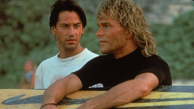 Keanu Reeves as Johnny Utah standing next to Patrick Swayze as Bodhi on the beach while he holds his surf board in Point Break (1991)