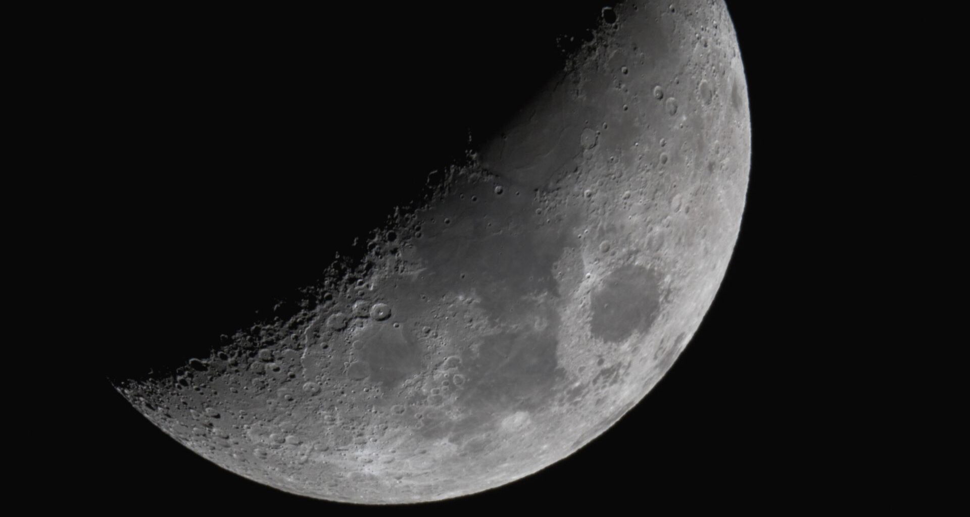The moon shines with its right half lit and its left veiled in shadow against a black night sky. Dark plains can be seen marking its surface around the equator, while large craters line the line separating night from day.