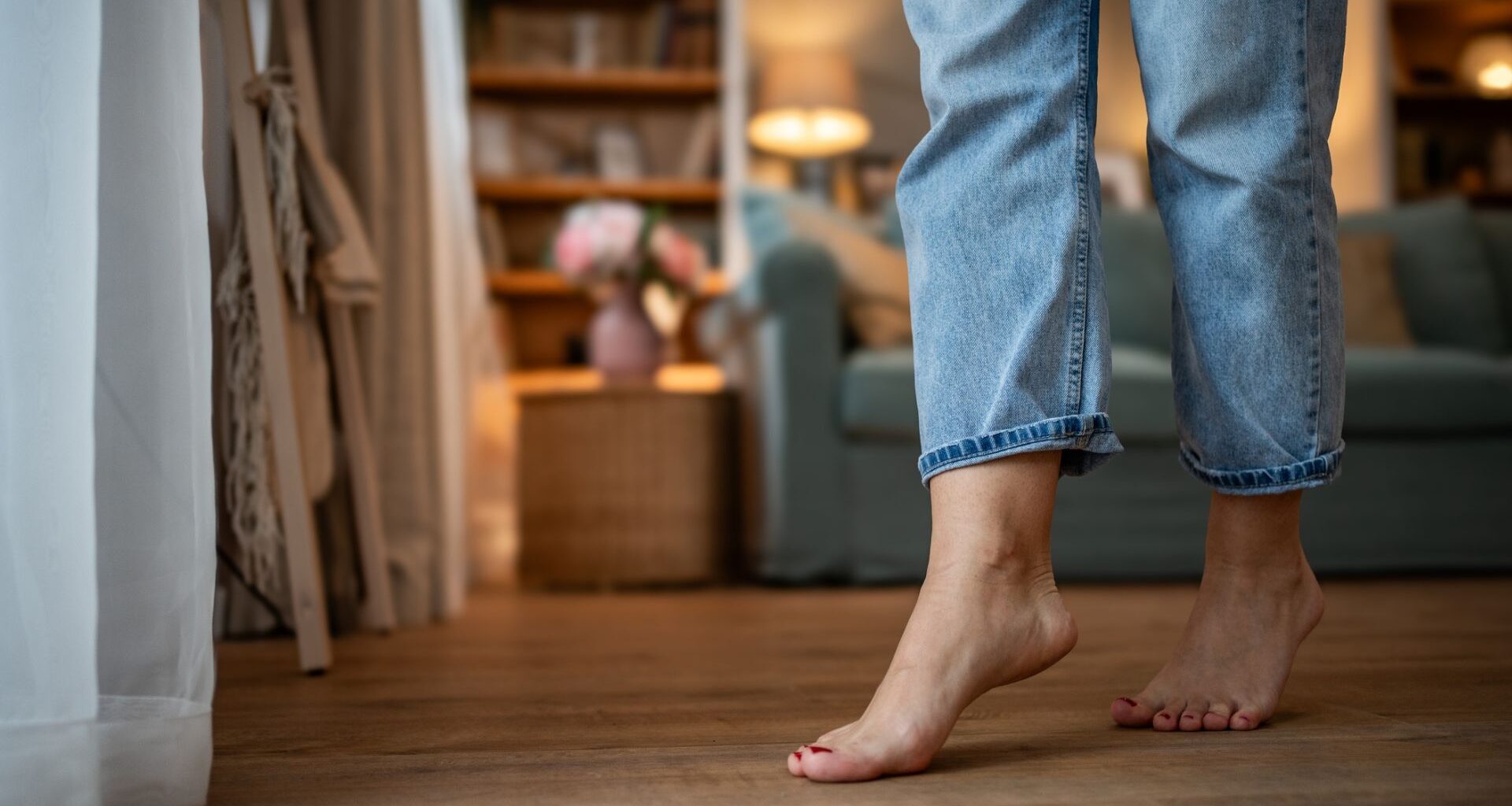 shot of bare feet in jeans on wooden floor with a living room scene blurred in the background. the feet are lightly on toes with red nails