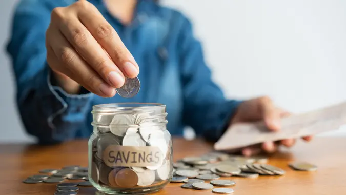 Woman hand is putting a coin into a glass bottle next to a pile of coins on a brown wooden table