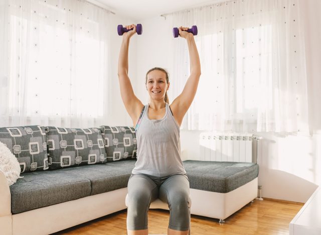 Smiling muscular sportswoman sitting on the chair in her apartment and doing fitness exercises for biceps with dumbbells.