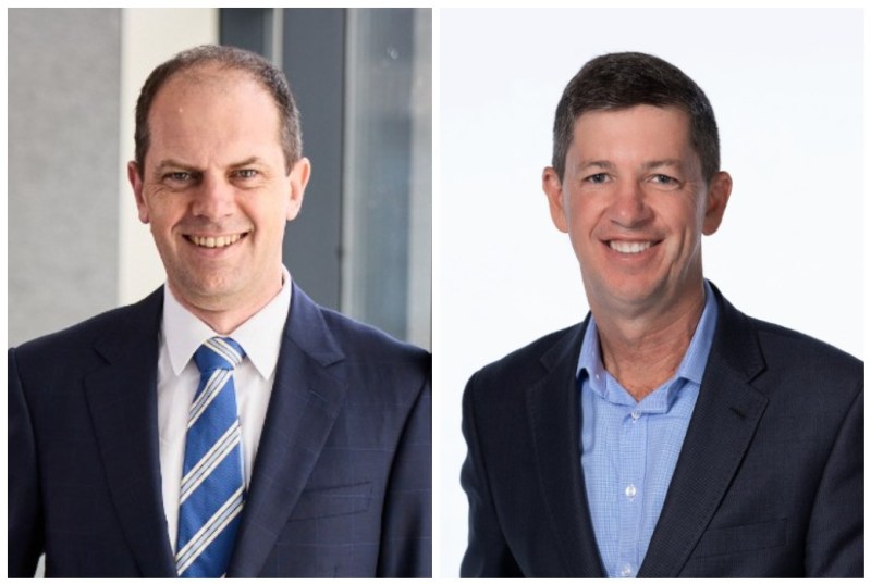 Two professional men smiling in business attire, one wearing a navy suit with a striped tie, and the other in a navy blazer over a light blue shirt. The background features modern office elements, highlighting a corporate environment.