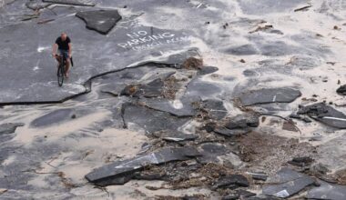 A man rides a bicycle across a coastal road that has been severely fractured and covered in sand and debris by a storm.