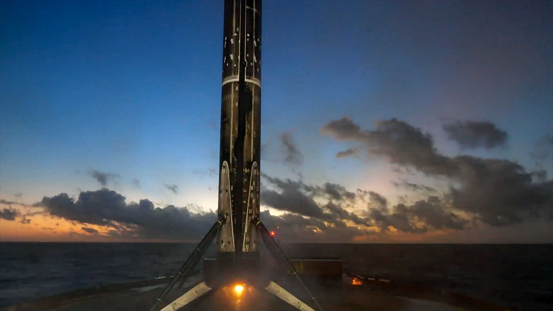 the first stage of an orbital rocket caked in black soot stands on its four landing legs atop an ocean-based droneship after a sunset propulsive landing from space