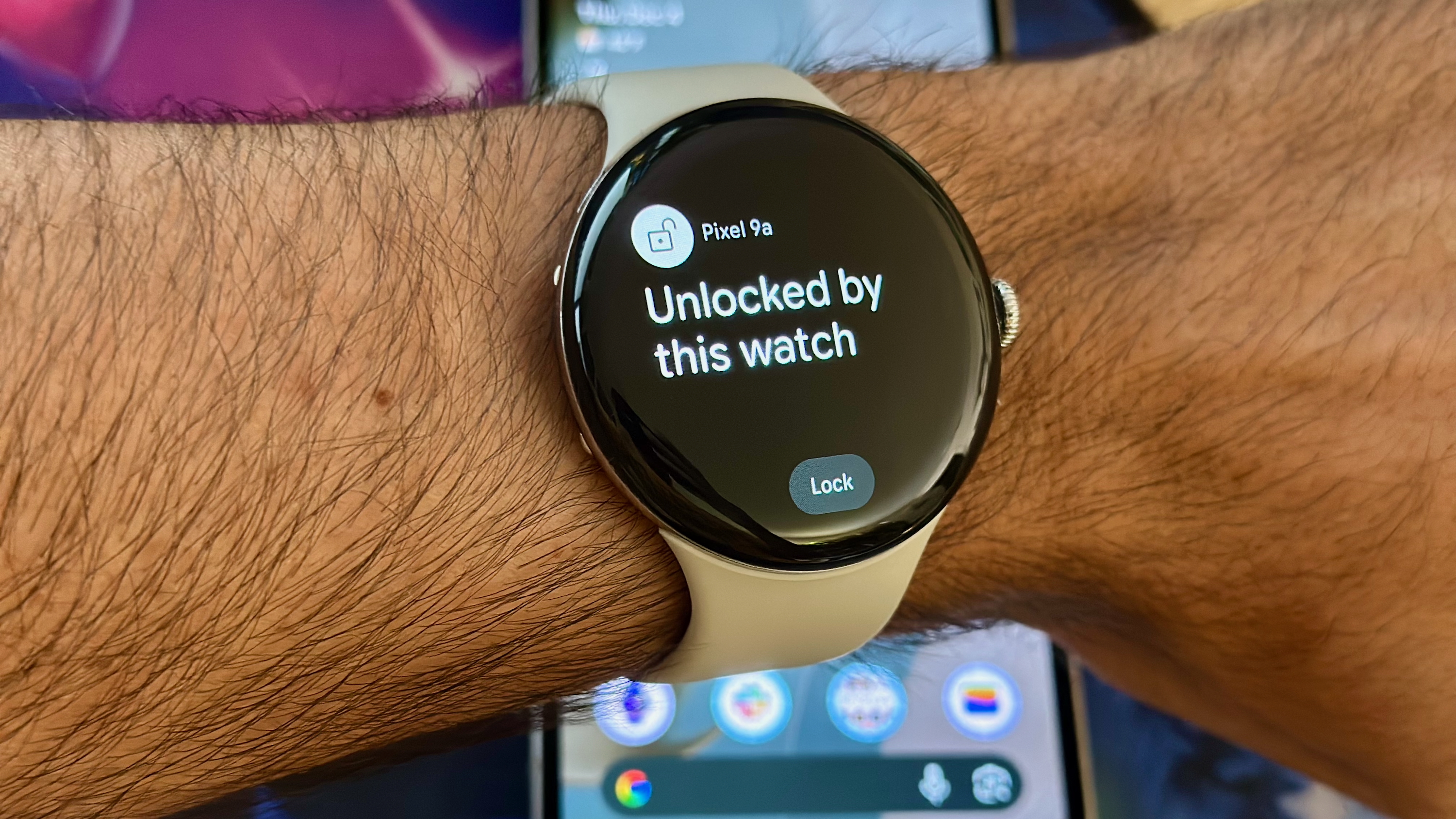 A photo of the Google Pixel Watch 4 worn above a Pixel 9a, showing the pop-up "Pixel 9a Unlocked by this watch" with a "Lock" button at the bottom.
