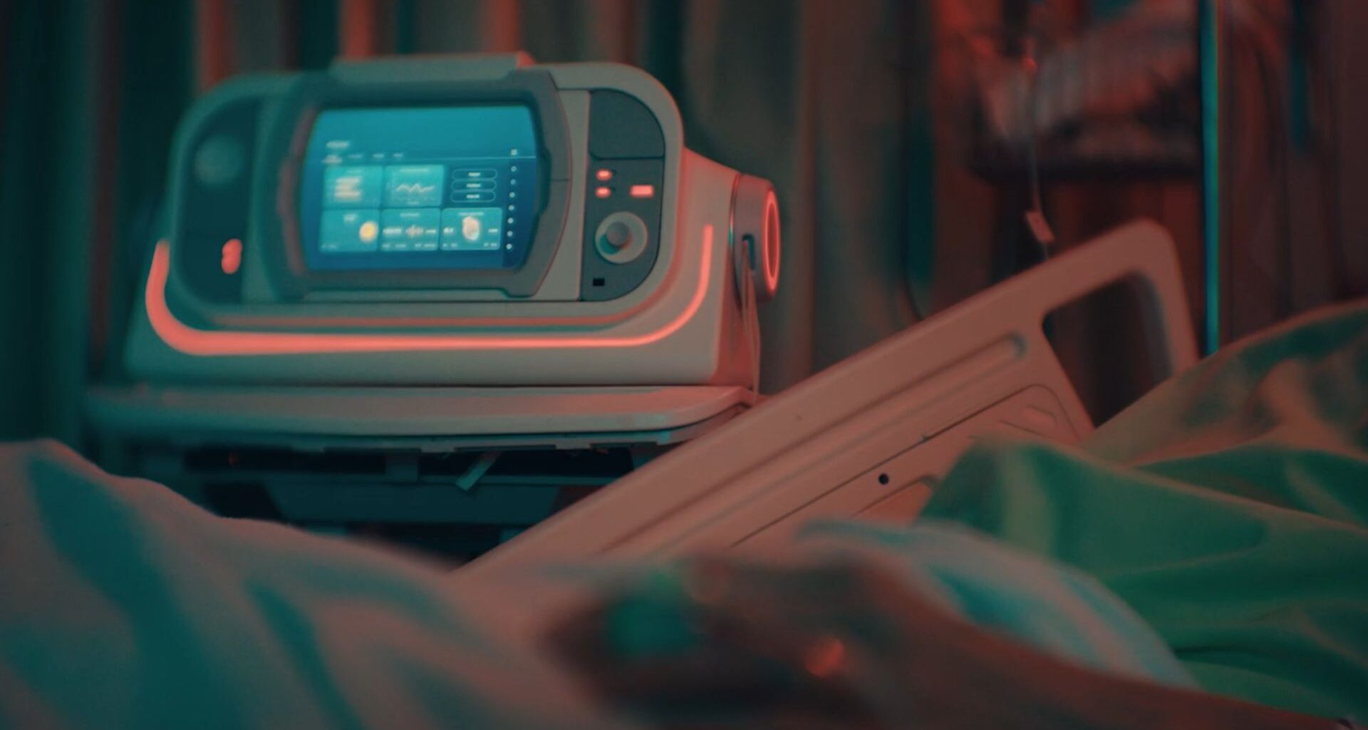 An image of a device with a screen and dials sitting next to a patient's bed in a hospital