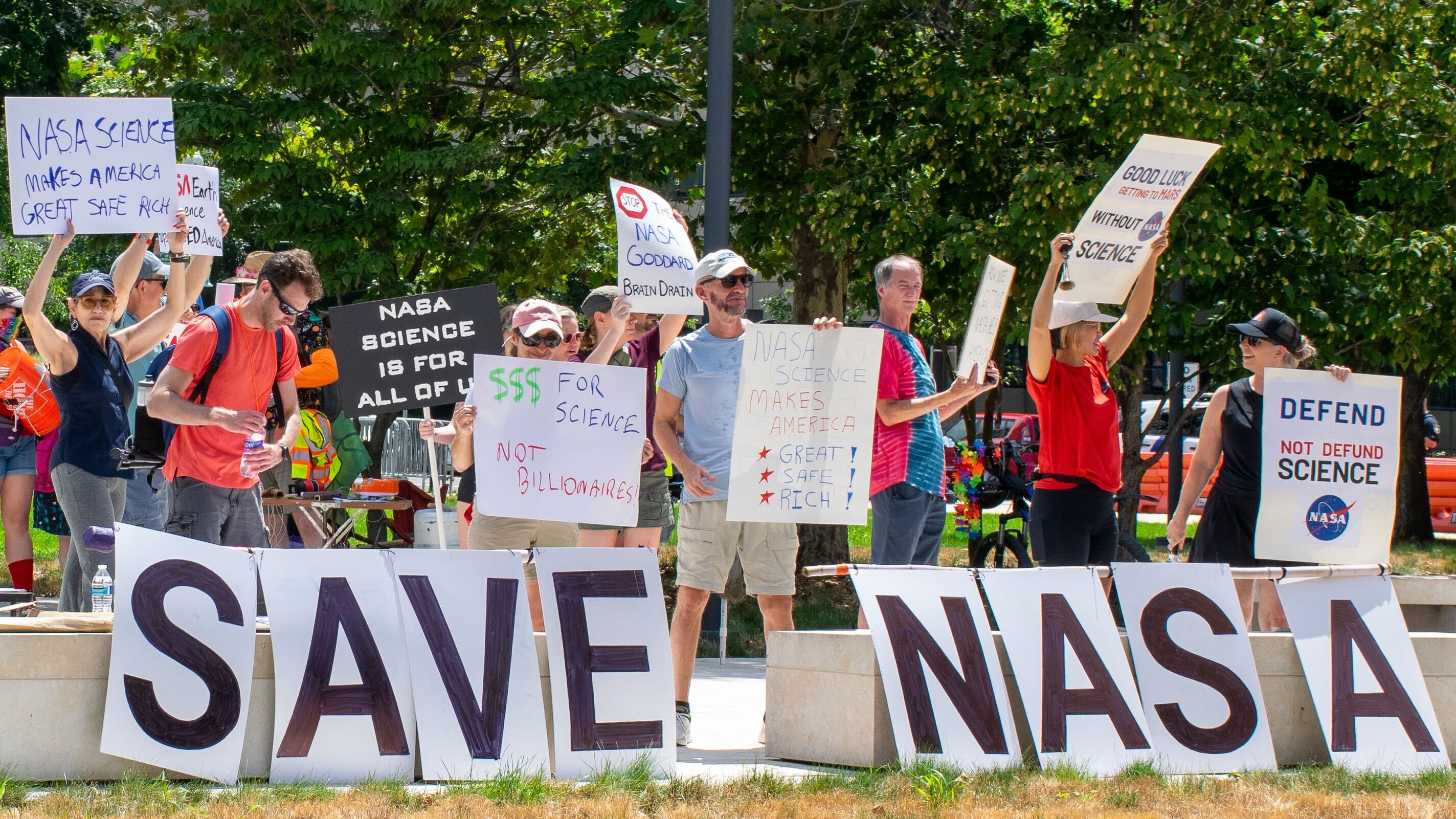 People hold signs outside on a sunny day in protest of NASA budget cuts, July 20, 2025.