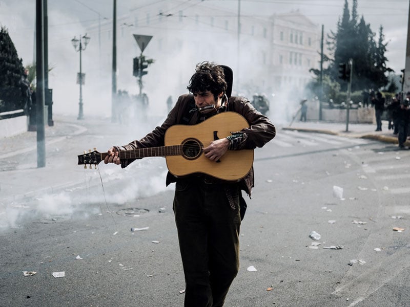 A man in a brown jacket plays an acoustic guitar while walking through a smoky street during a protest, with debris scattered on the ground and people in the background.