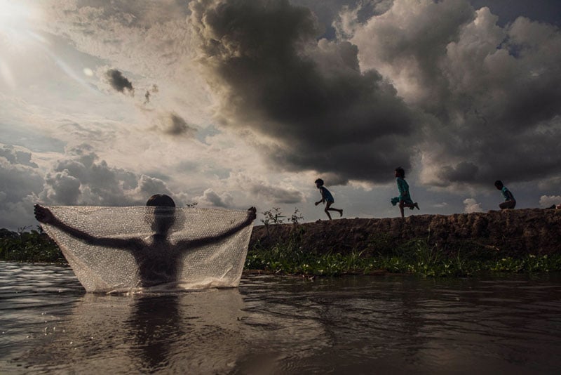A silhouetted child stands in water holding up a fishing net, while three children run along a grassy embankment under a dramatic, cloudy sky. Sunlight breaks through the clouds, casting reflections on the water.