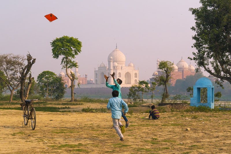 Children fly a red kite in an open, sandy area with scattered trees. The iconic white Taj Mahal stands in the background, and a bicycle and small blue structure are visible nearby. The sky is hazy and soft.