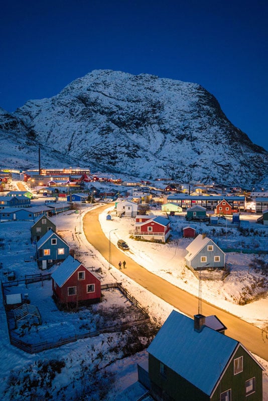 A snowy village at dusk, with colorful houses lining a winding road illuminated by streetlights, set against a backdrop of a large, snow-covered mountain.