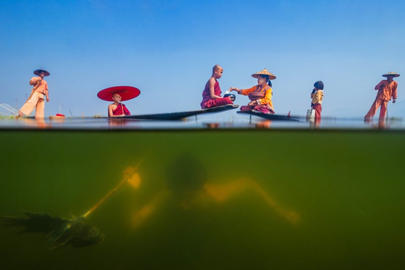 A split-level photo shows monks in traditional robes on a boat above water, with one monk paddling underwater. The scene is bright and colorful, with a clear blue sky and reflections on the water’s surface.