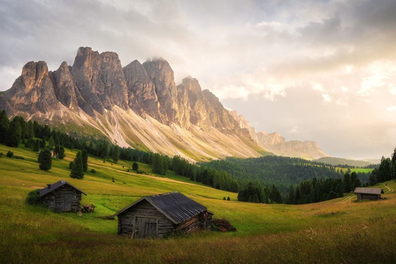 Three wooden cabins sit on a grassy hillside with tall, rugged mountains and a forest in the background, illuminated by soft, golden sunlight under a partly cloudy sky.