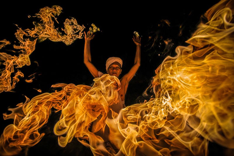A person wearing a white headscarf raises their arms amid large swirling flames against a dark background, creating a dramatic and intense scene.