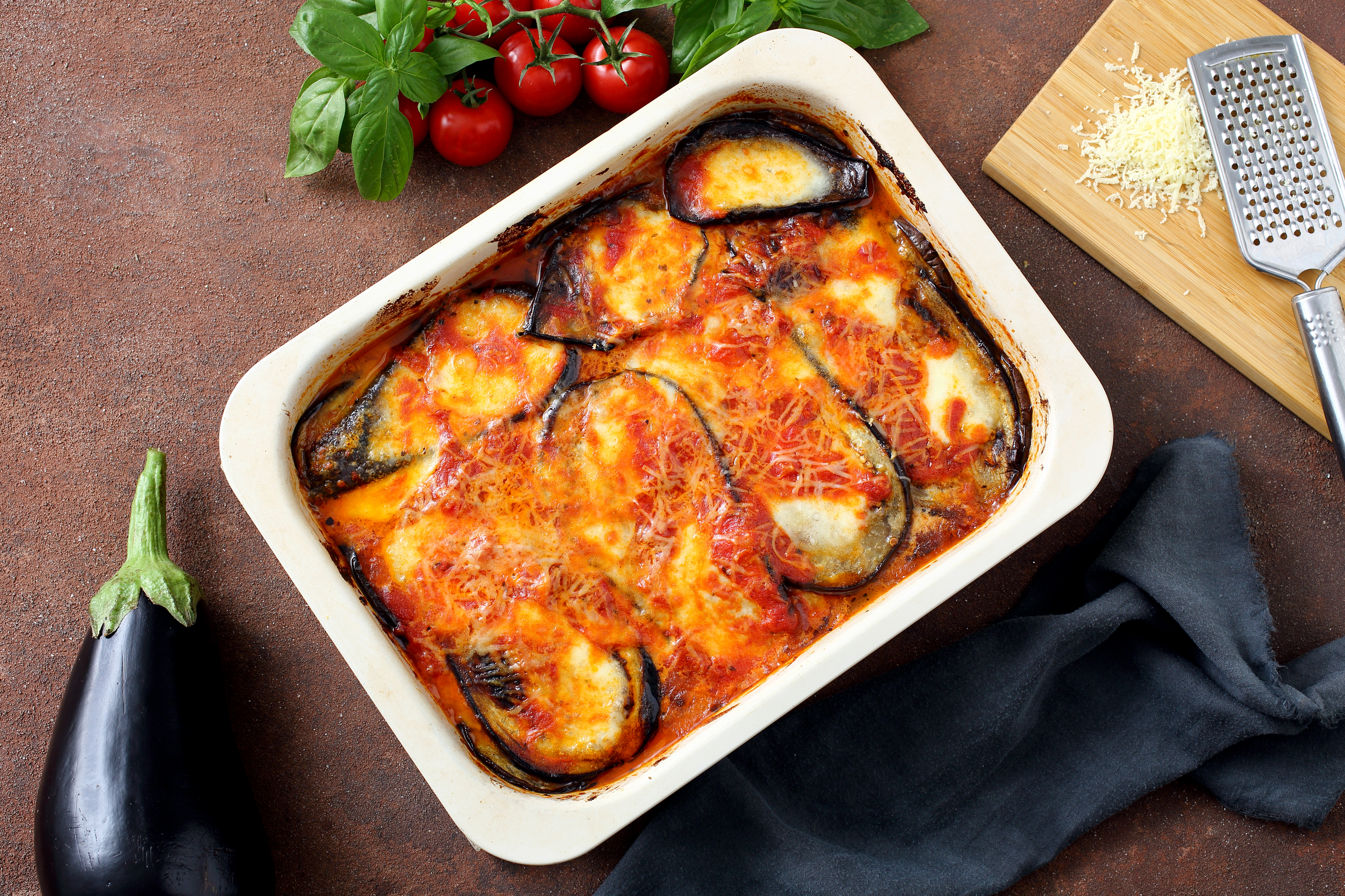 Eggplant Parmigiana in a baking dish, with a raw eggplant, basil, cherry tomatoes, and grated cheese with a grater on the side.