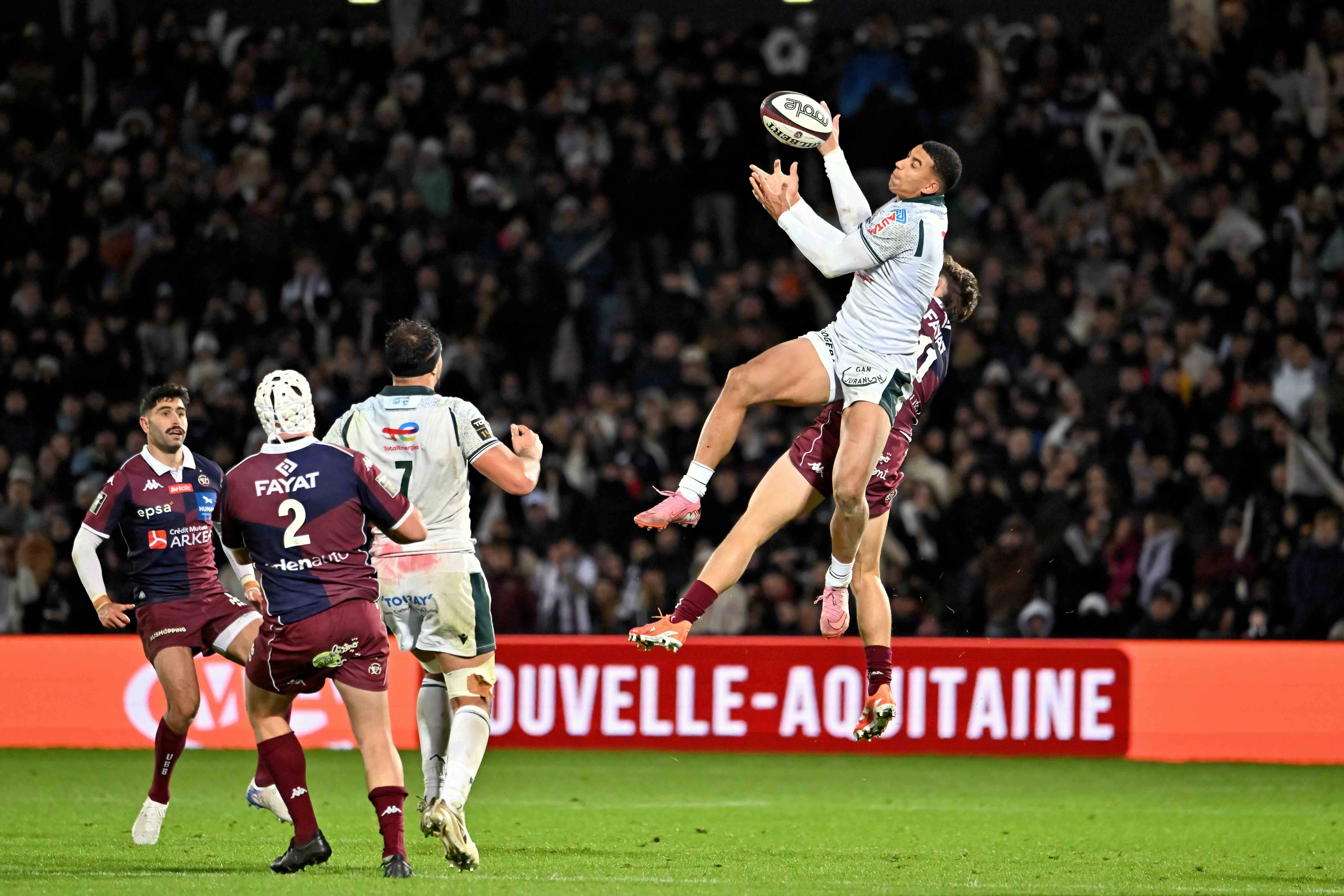 Aaron Grandidier-Nkanang of Pau (white jersey) catching a rugby ball mid-air against Xan Mousques of UBB (maroon jersey) during a Top 14 rugby match.